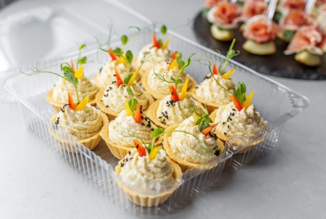 Assorted savory tartlets with piped cheese or egg filling, garnished with herbs and peppers, presented in a clear plastic container.